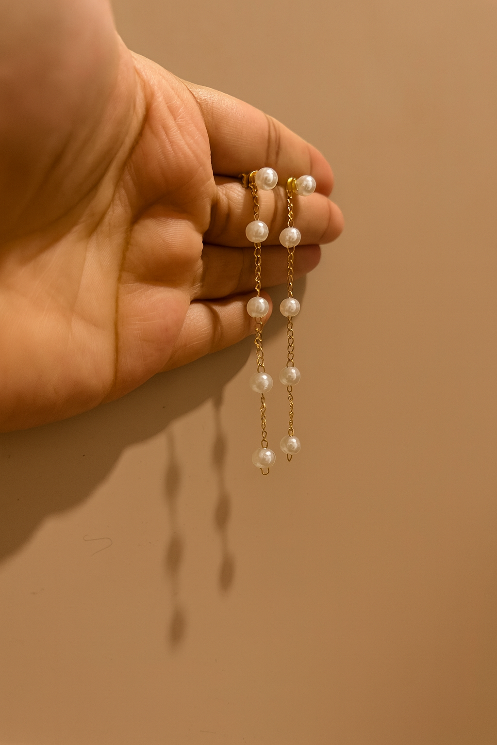 Pearl drop earrings held between fingers against a beige background