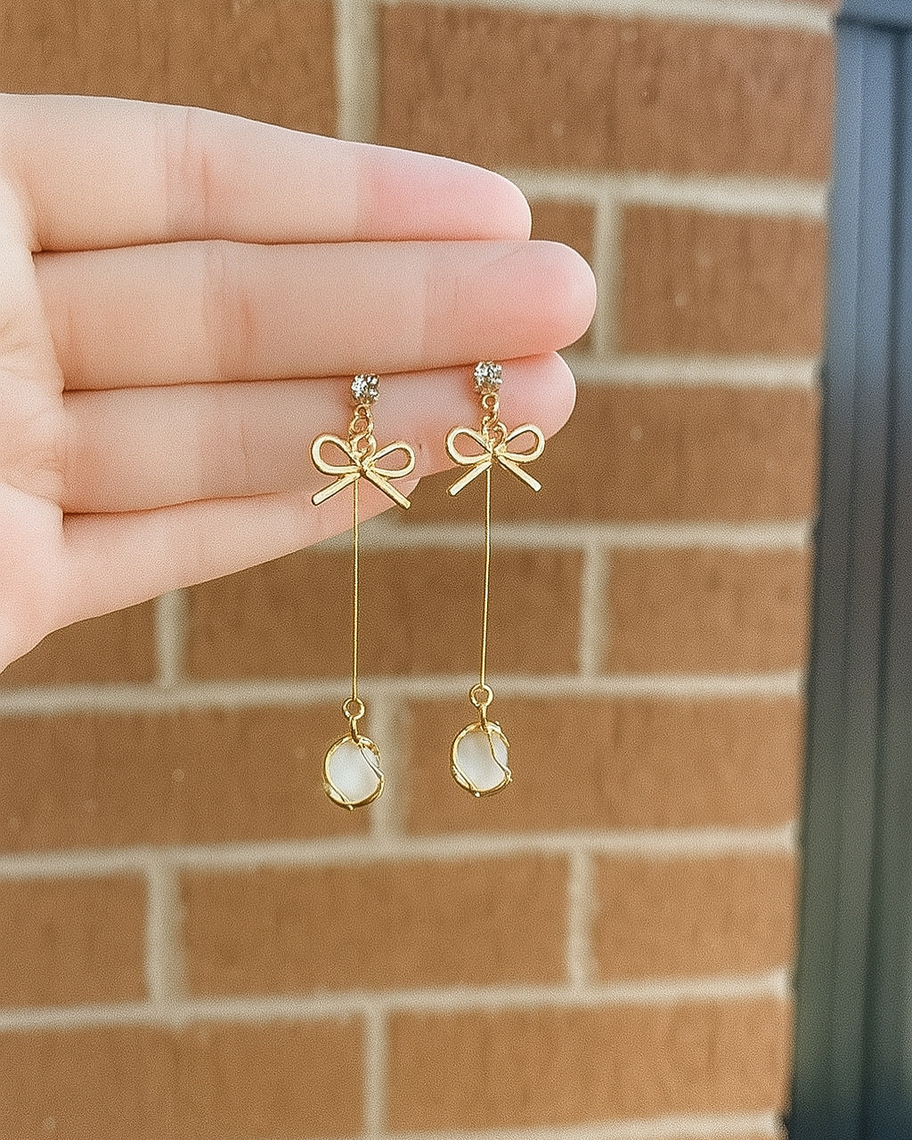 Gold bow-shaped earrings with dangling elements held by a hand against a brick wall.