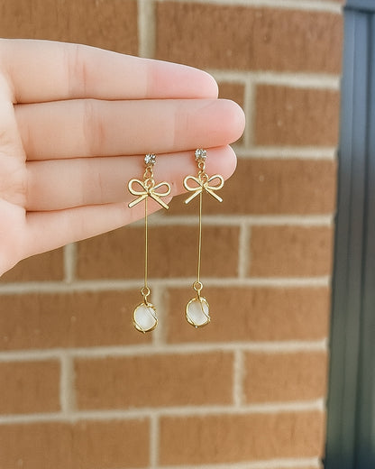 Gold bow-shaped earrings with dangling elements held by a hand against a brick wall.