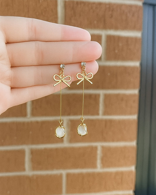 Gold bow-shaped earrings with dangling elements held by a hand against a brick wall.